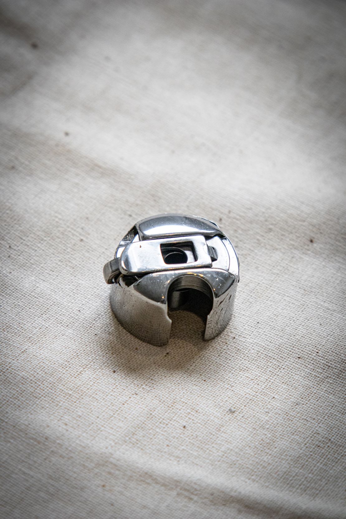 Angled top-rear view of the RMIT Store bobbin case, revealing the wide rear slot and detailed chrome finish, photographed on neutral beige cloth background for texture contrast.